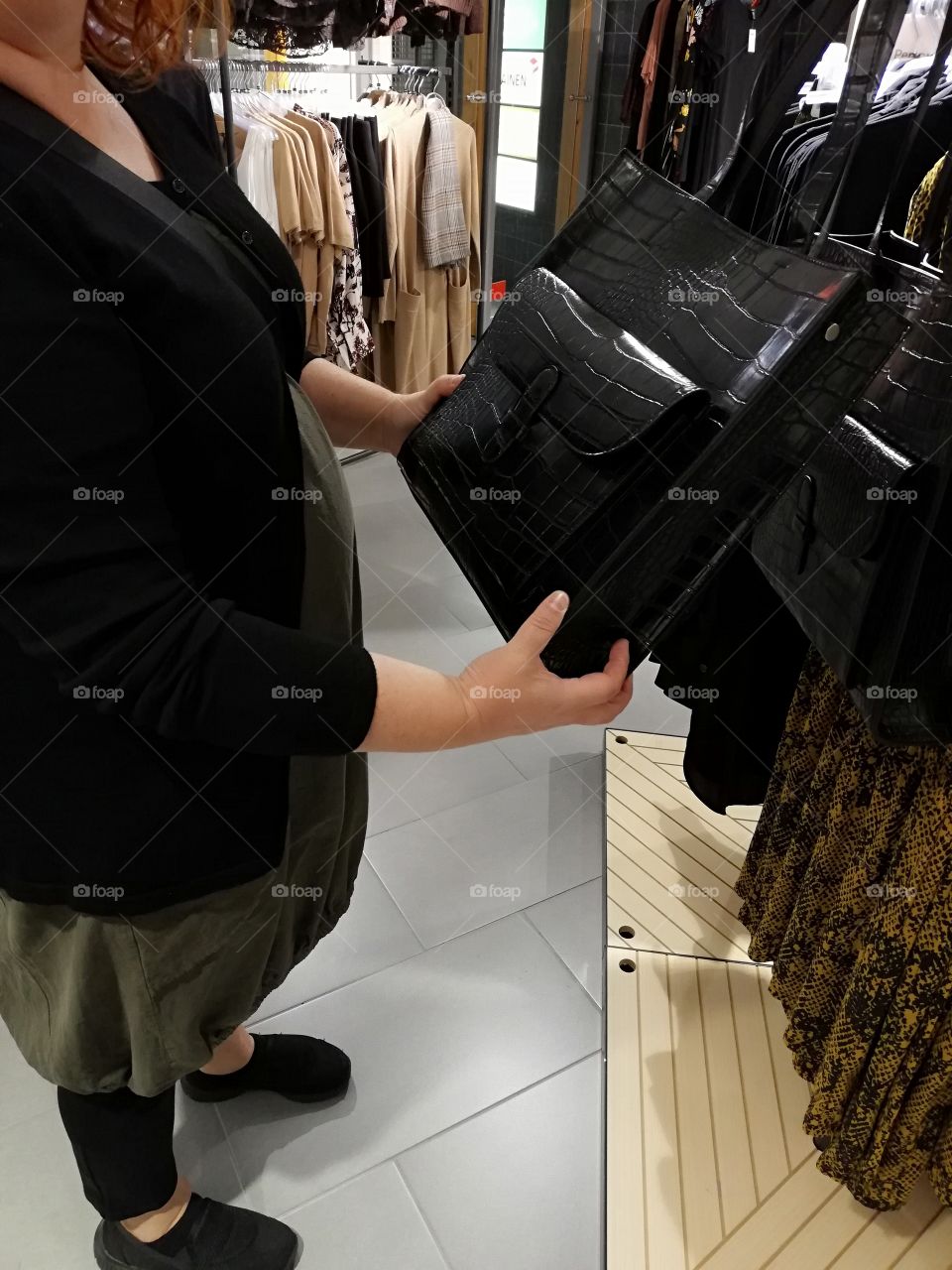 A red haired woman regarding a new fashion handbag. A black glossy bag hanging among the others. In the background the clothes on the hangers and a checkered scarf around the collar of the coat.