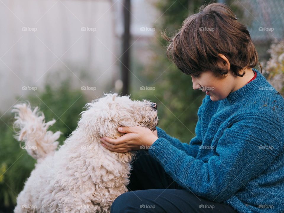 Cute little boy with dark hair wearing blue knitted sweater with white curly poodle dog in autumn garden, portrait of child 