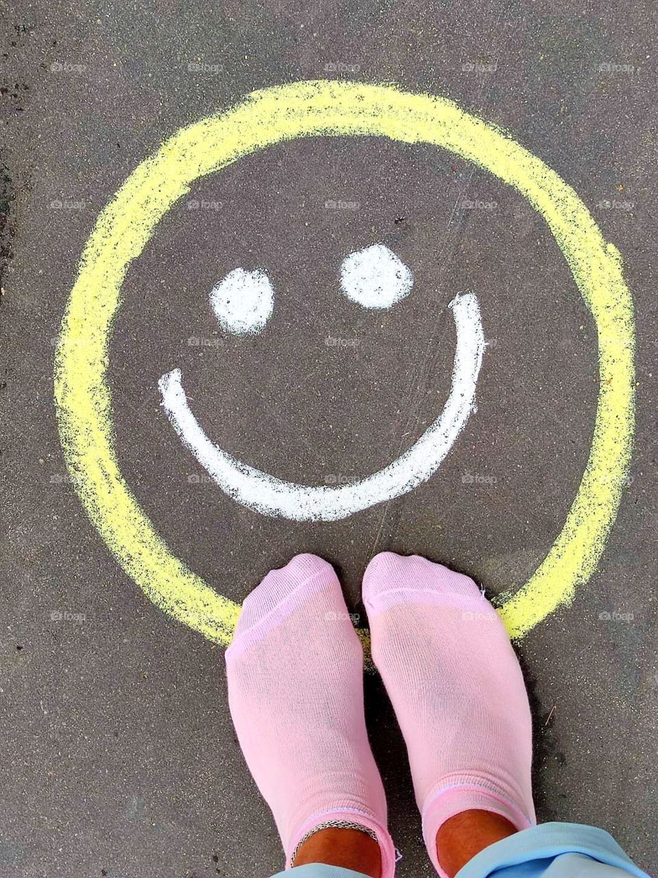 A smiling emoticon is drawn on the pavement.  Feet in pink socks stand on a smiley face