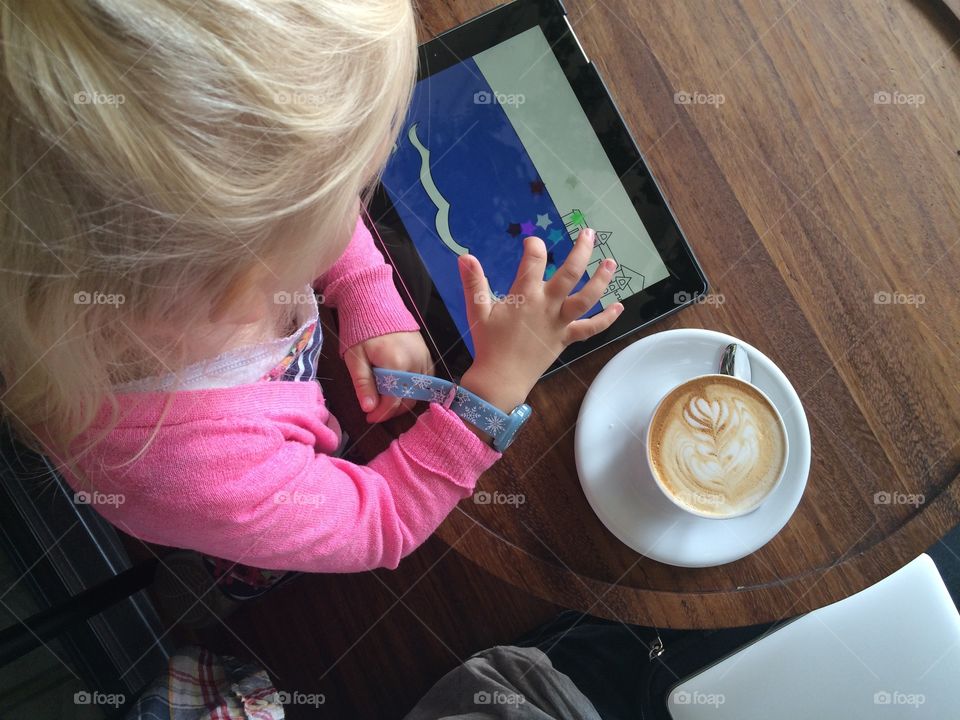 Little girl drinking hot chocolate and playing on an iPad.