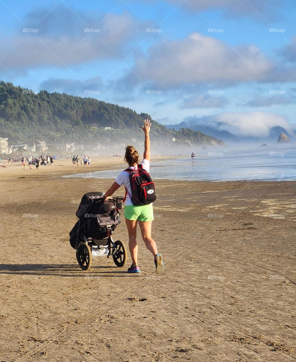 young mother wearing a backpack pushing a stroller waves to her family down the beach on a Summer evening in Oregon