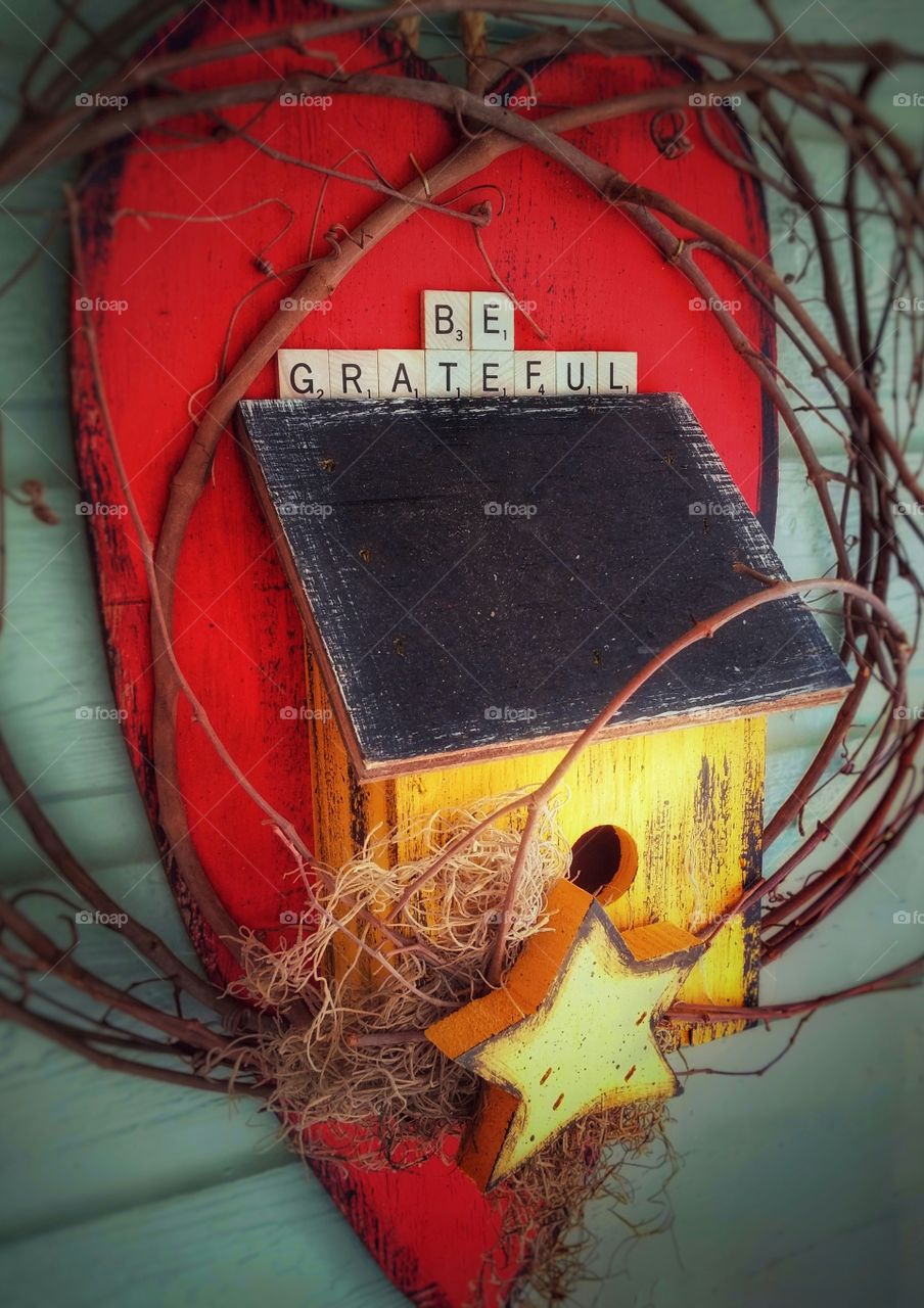 Pop of a Red heart behind a yellow birdhouse hanging on green siding with Be Grateful written on it