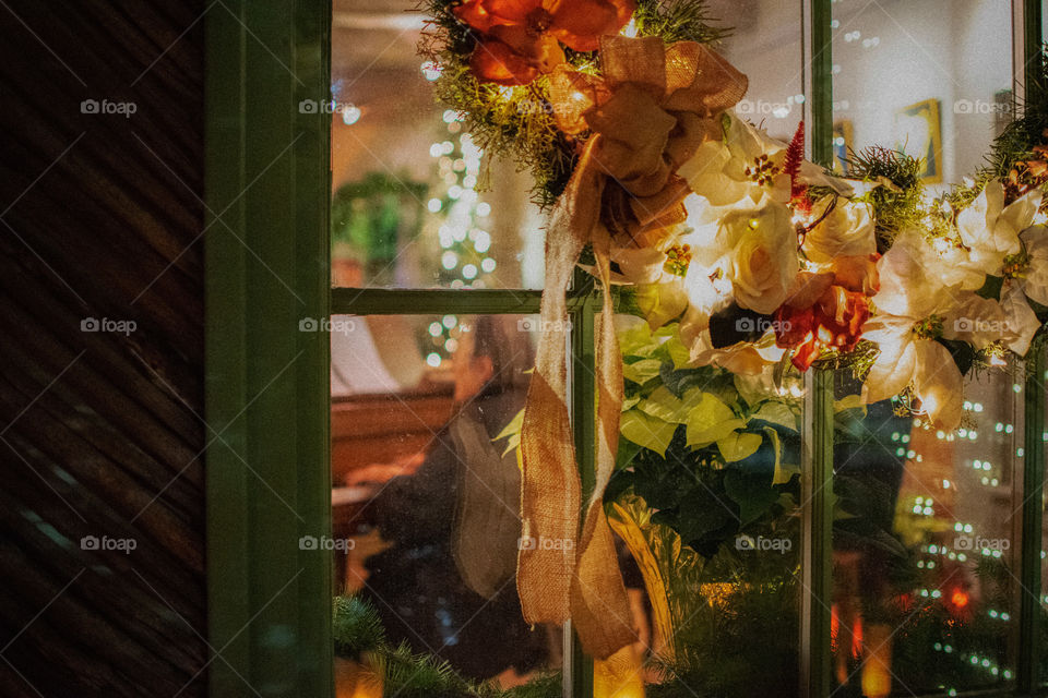 Christmas wreath on the glass window of a house with an elderly woman playing piano seen through the glass 