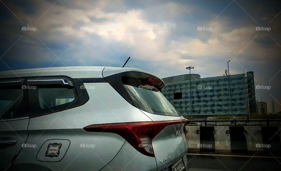 This dynamic photo captures a car racing down the highway, with half of the vehicle in frame and buildings and the sky providing a dramatic background. The sense of motion and energy is palpable.
