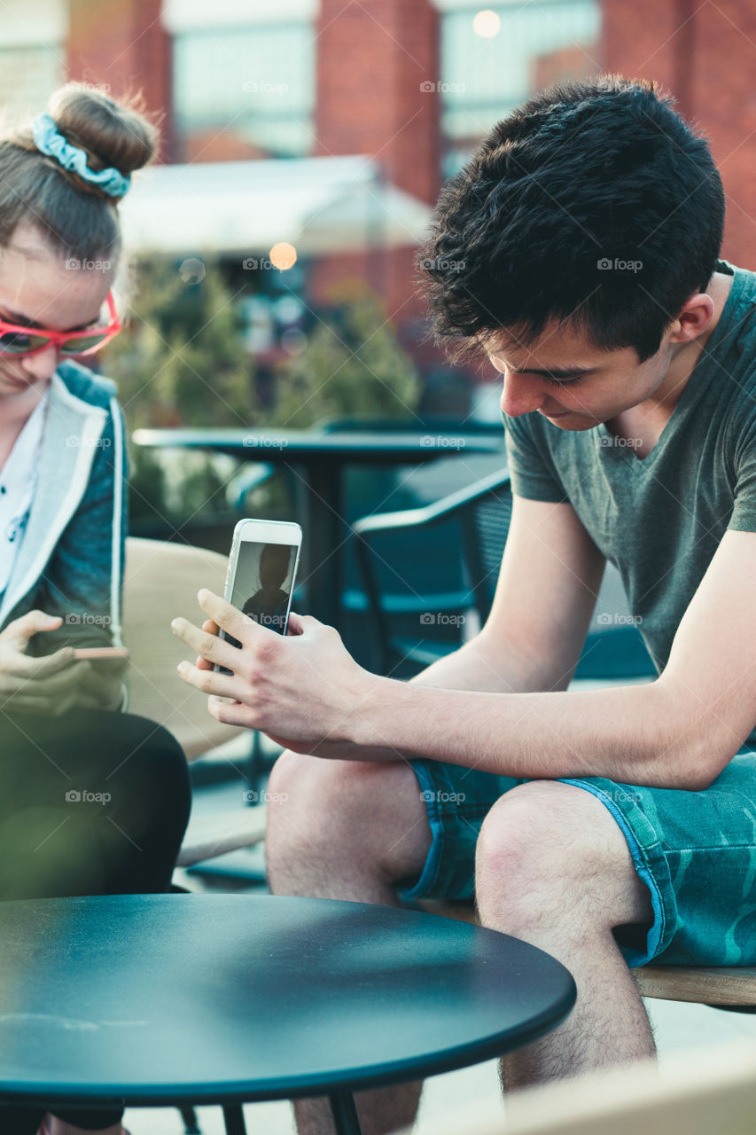 Couple of friends, teenage girl and boy, having fun using smartphones sitting in center of town, spending time together