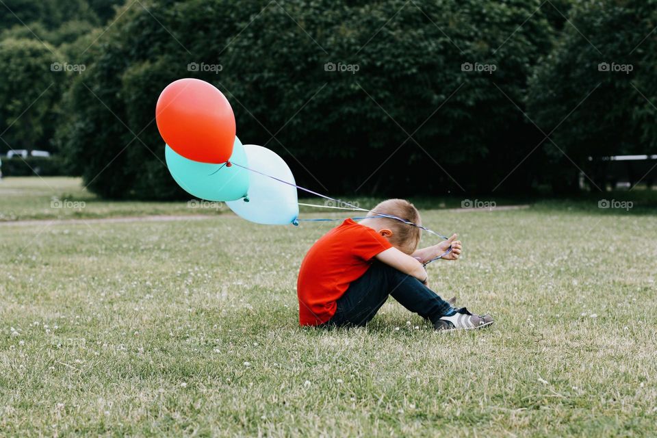 A little blond boy in a red T-shirt sits on a green field, holding a bunch of colorful balloons in his hand, side view 