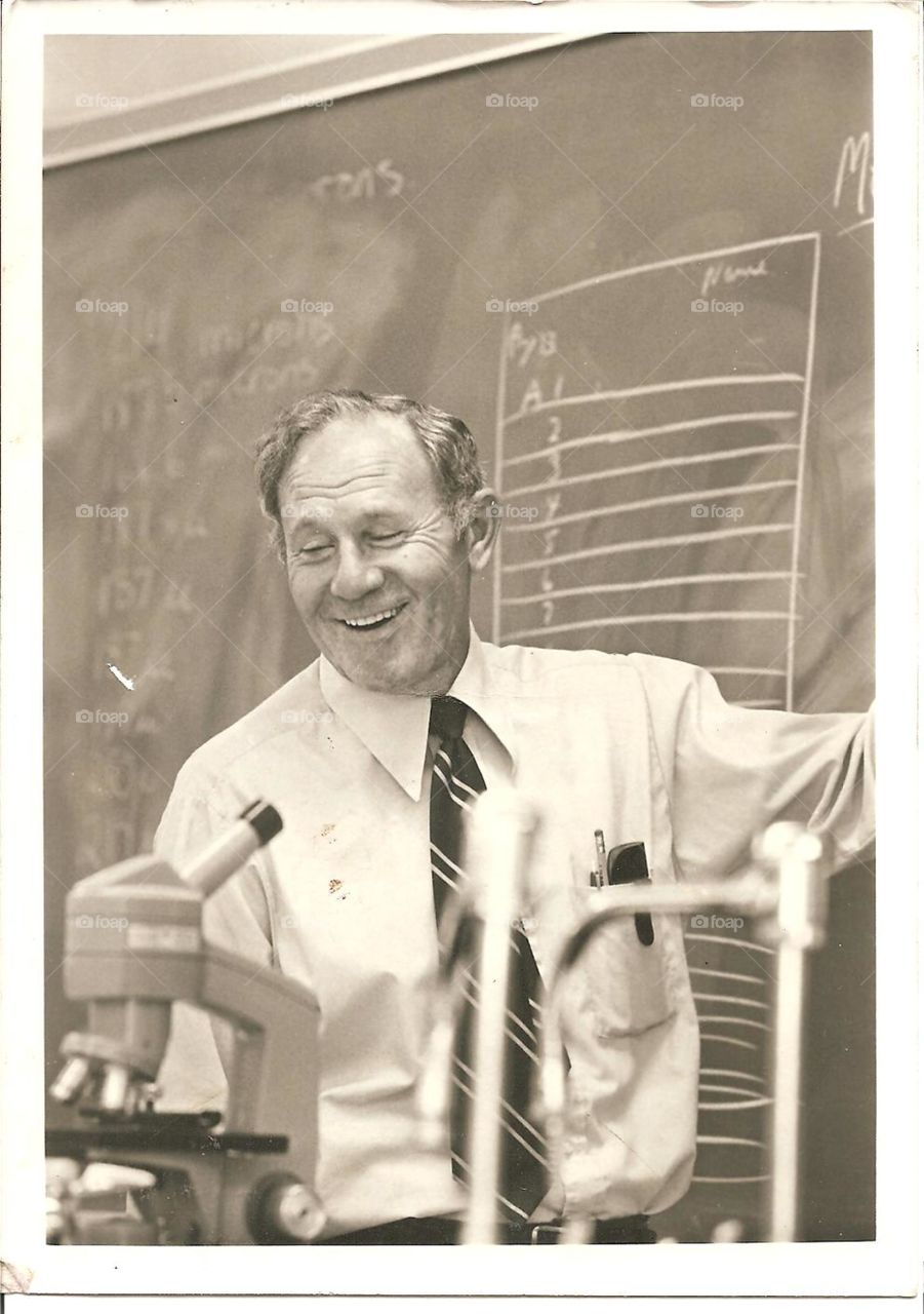 B&W vintage candid 1960s pic of biology high school teacher in class, smiling at blackboard near microscope.