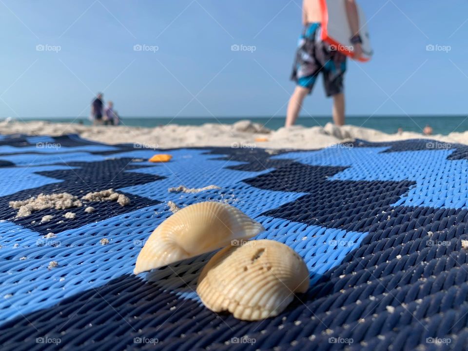 Ready for surfing or body board swim in the Atlantic Ocean at the beach in a nice vacation time with seashells on a blue sand proof carpet with people walking on the shore.