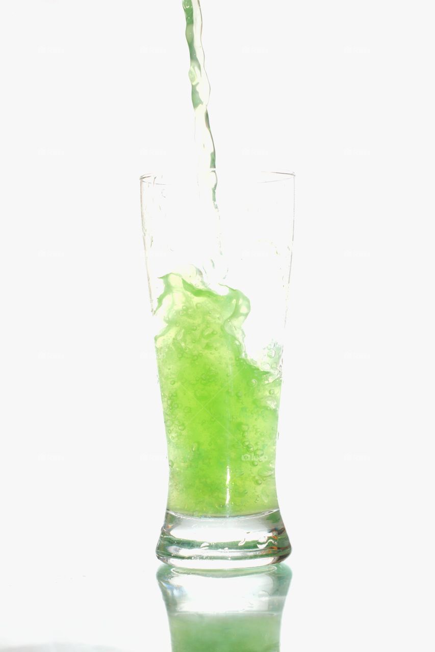 Líquido verde caindo em um copo de vidro com um fundo Branco e superfície branca.