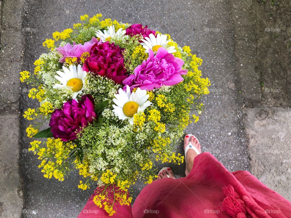 Woman in pink dress holding a giant bouquet of summer flowers consisting of pink peonies, daisies and yellow goldenrod