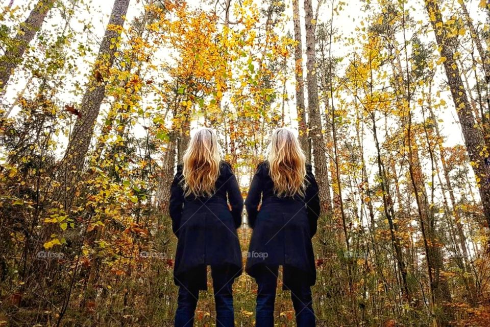 Two women with black coats and blonde hair stand in an autumn forest