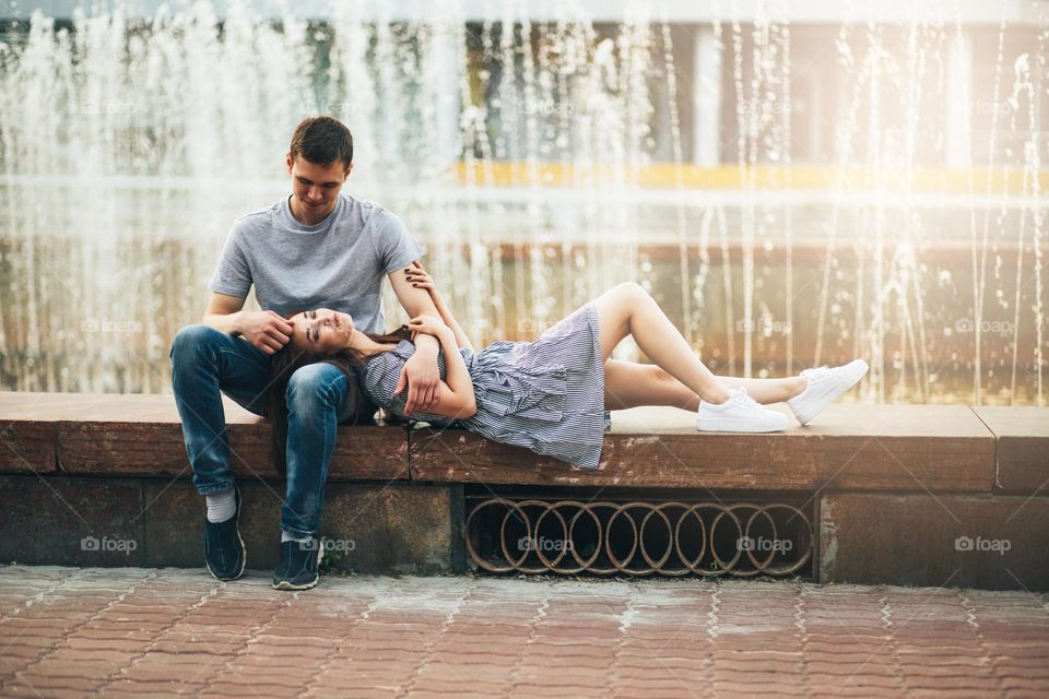 Happy young couple in love teenagers friends dressed in casual style sitting together near fountain on the city street