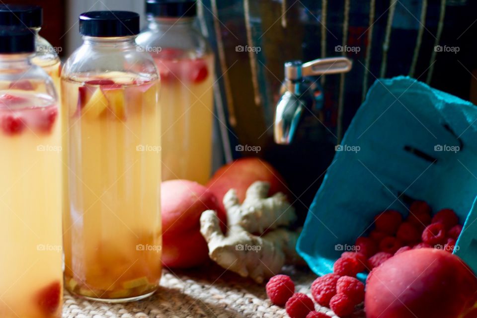 Kombucha, bottled for a second-ferment, flavored with nectarines, raspberries and ginger root slices on a natural fiber mat, whole nectarines and raspberries in a paper carton, stoneware kombucha crock with a stainless steel spigot in the background