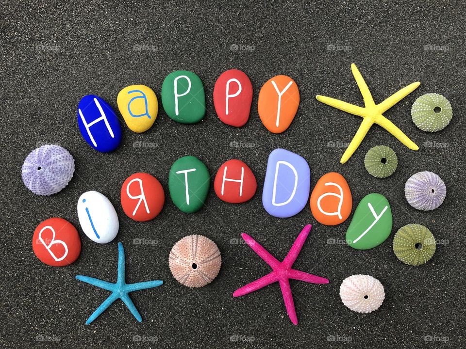 Happy Birthday with a funny composition of colored stones, starfishes and sea urchin shells over black volcanic sand
