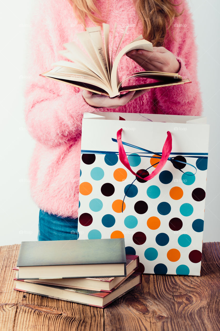 Young girl turning the pages of book in bookstore. Putting the books into paper bag. A few books on a wooden table. Teenager girl wearing pink sweater and blue jeans. Vertical photo