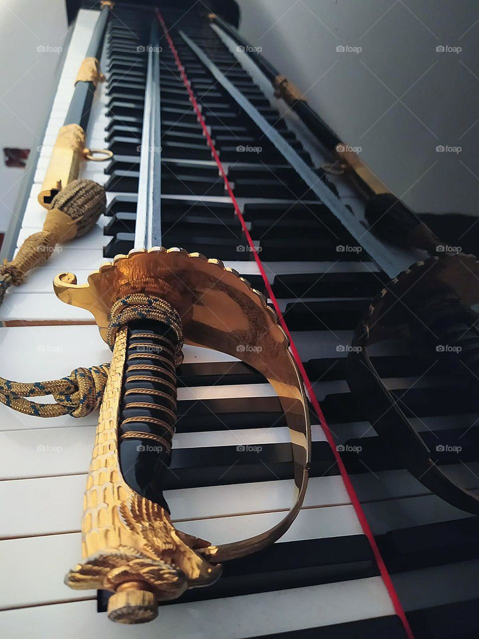 On the wooden keys of the piano lie a metal sword and scabbard. The sword consists of a metal handle with a bird's head and a metal blade. The sword reflects off the black wooden surface of the piano. Ceremonial sword of Italian military aviatio