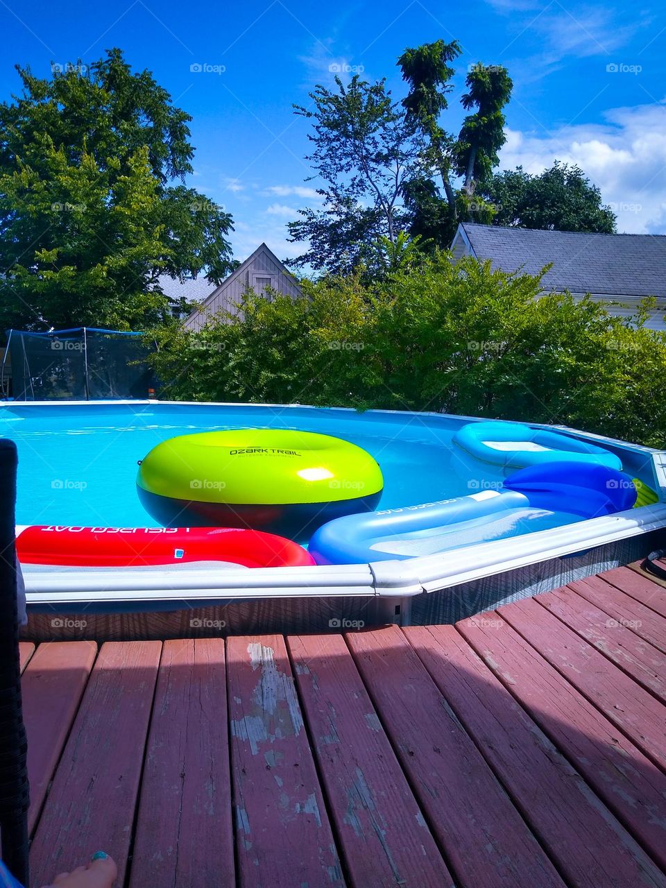 Back yard pool with summertime vibes bright blue water and sky’s with floating pool toys and a rustic back porch. 