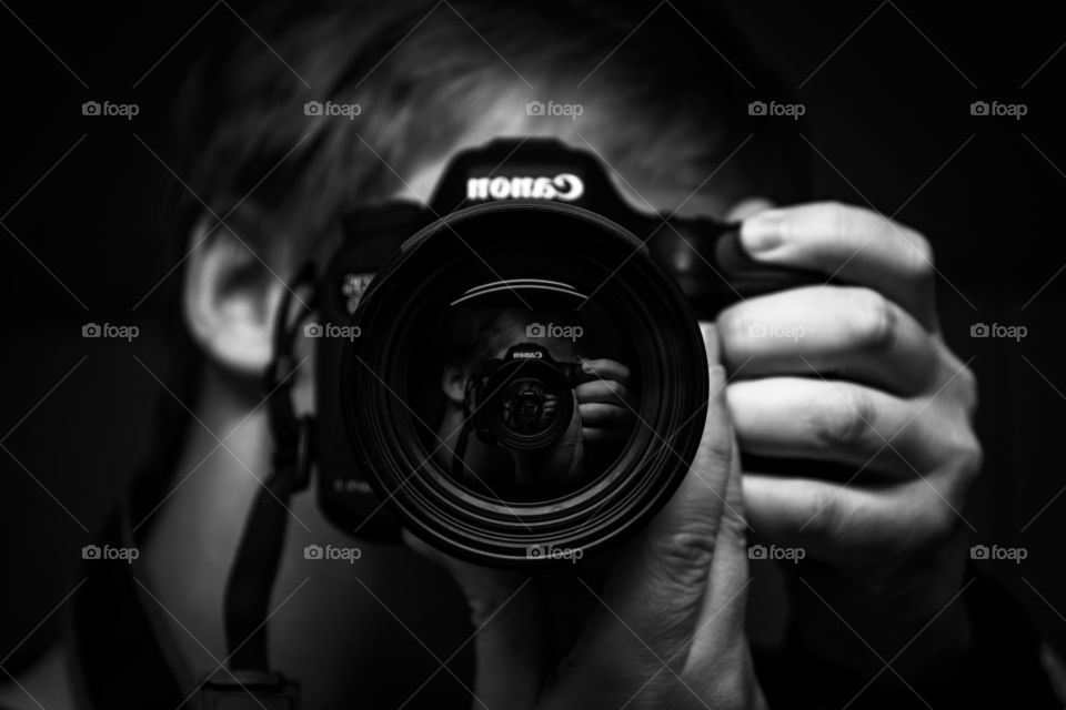 A black and white portrait of a photographer taking a selfie in a horror holding his dslr camera in front of him with infinite reflections inside of the camera's lens.