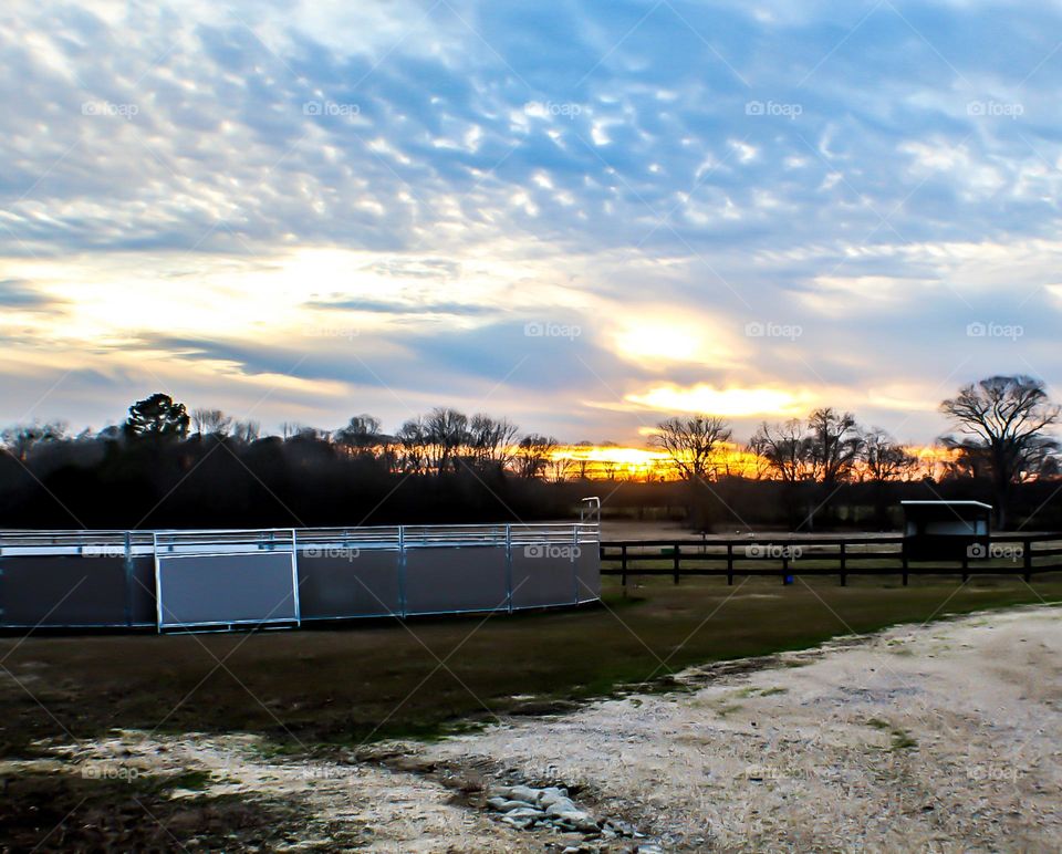 The end of a colorful sunset at a horse ranch. Wispy clouds and trees are seen on the horizon.