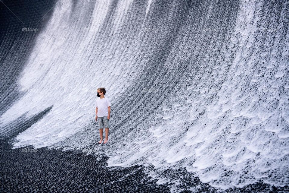 A boy in denim shorts and a white T-shirt looks at an artificial waterfall. Creative fountain in the form of a waterfall. Minimalistic photo in gray tones. Conceptual water installation. Expo 2020 Dubai