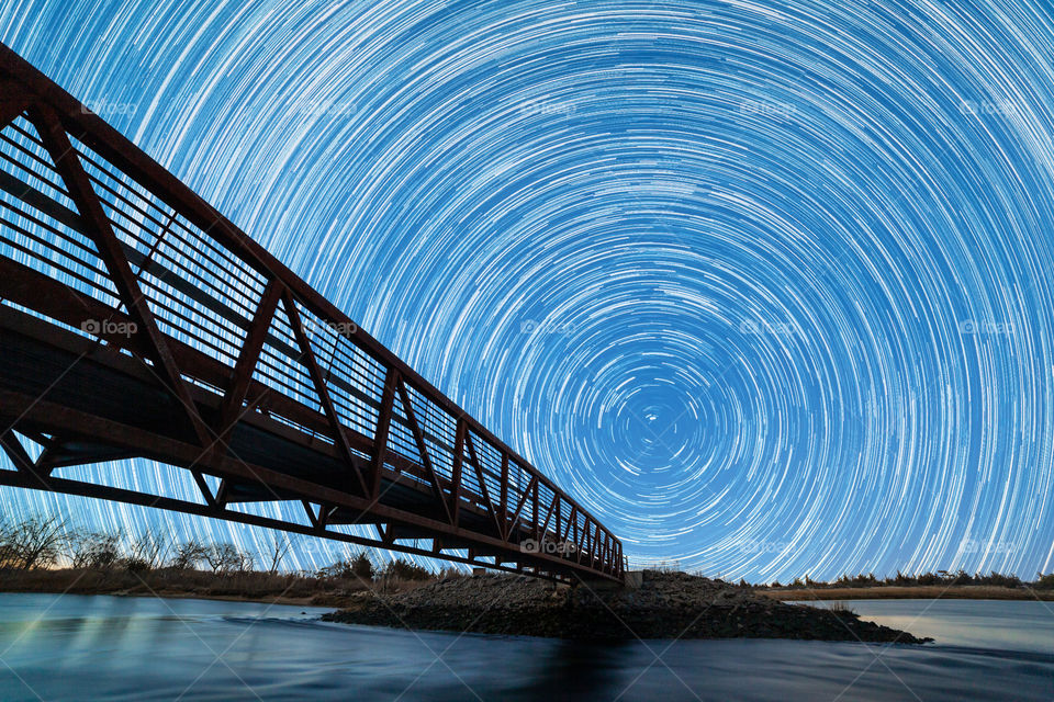 Silhouette of a footbridge crossing a stream of water, with circular star trails in the night sky and Polaris (the North Star) just above the horizon. 