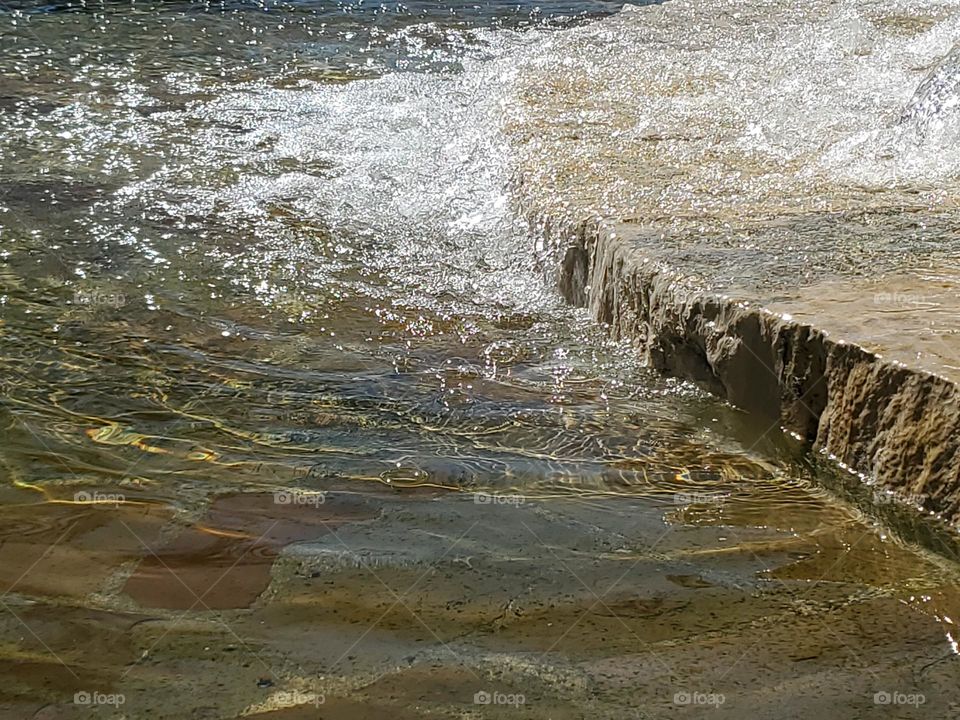 Closeup of a water fountain that fills an urban Spring fed pool with water at a city park. The surface appears to be made out of flagstone and local limestone.