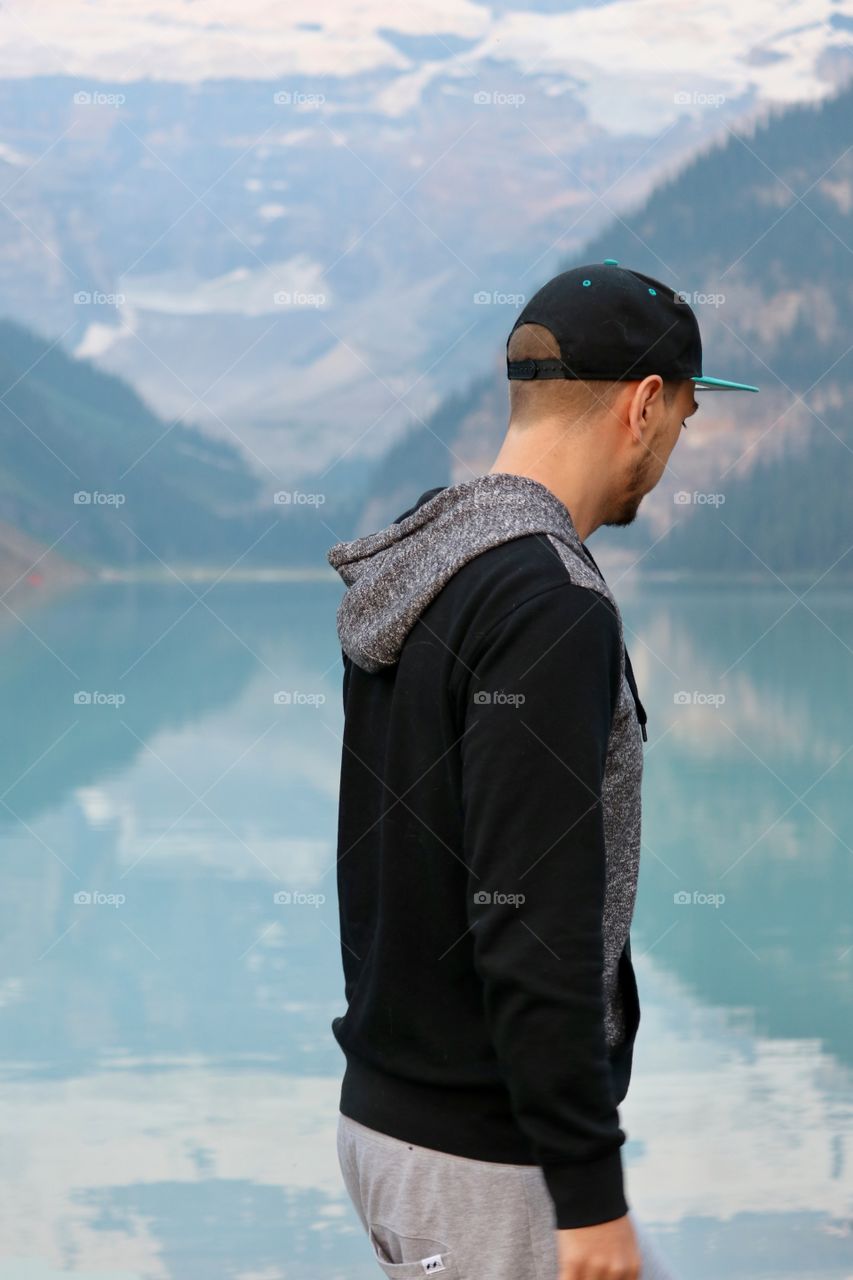 Side profile young man lost in thought wearing baseball cap and black hoodie standing in front of scenic turquoise glacial lake and mountains at lake Louise in Canada’s Rocky Mountains 