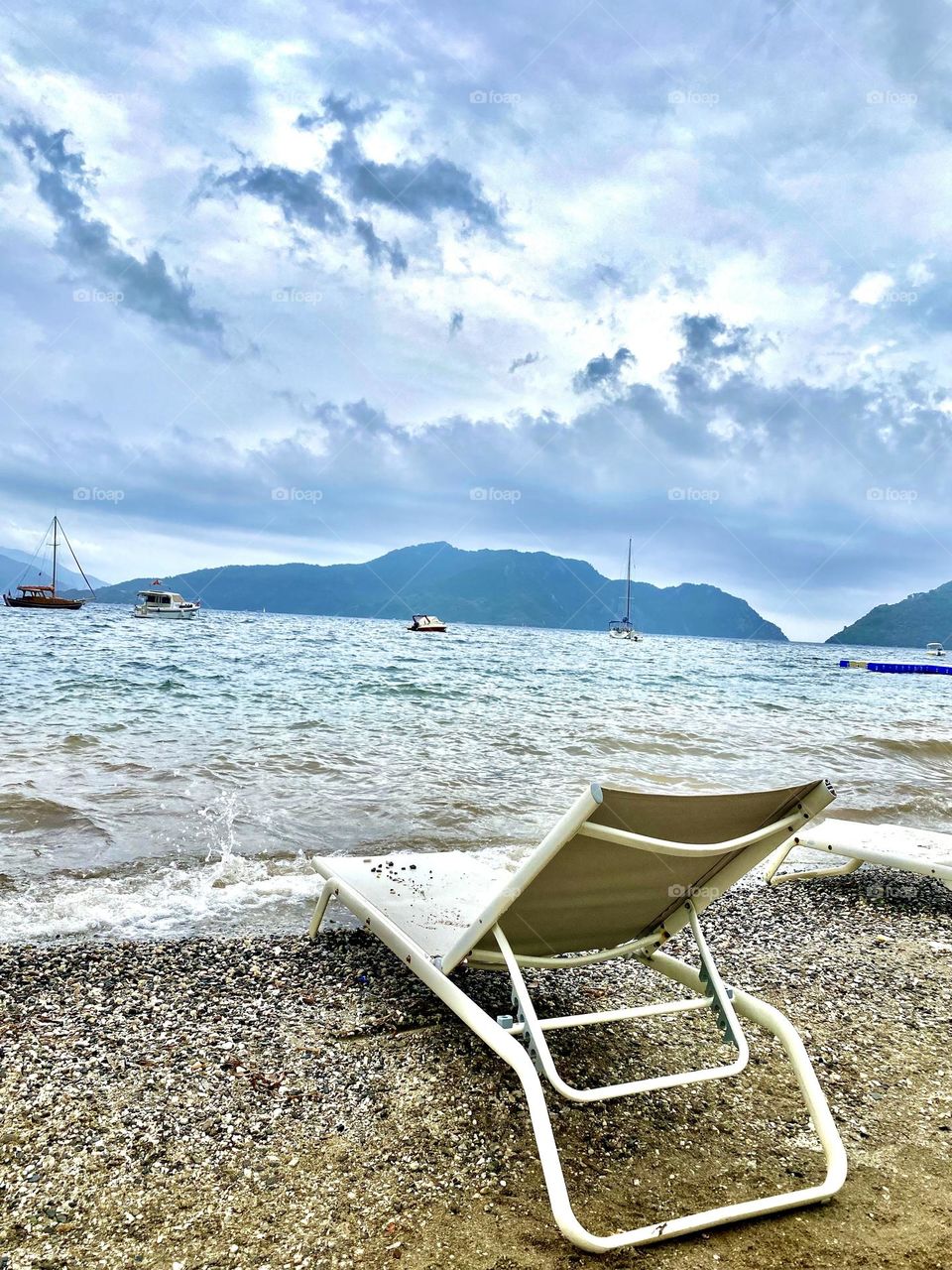На фотографии изображен лежак, расположенный на песчаном пляже у самого берега моря. Вдали, на спокойной морской глади, видны несколько небольших корабликов, которые покачиваются на волнах.