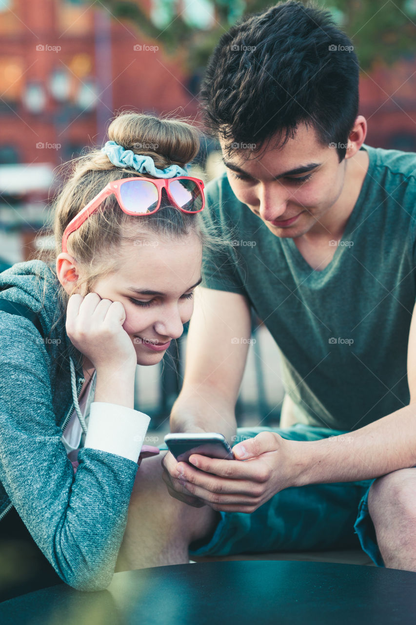 Couple of friends, teenage girl and boy, having fun with smartphones, sitting in center of town, spending time together