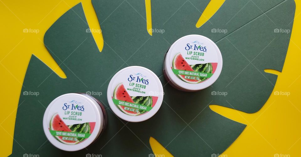 Three St. Ives juicy watermelon lip scrubs diagnolly set on a green paper leaf with a yellow background. (Facebook ad size)