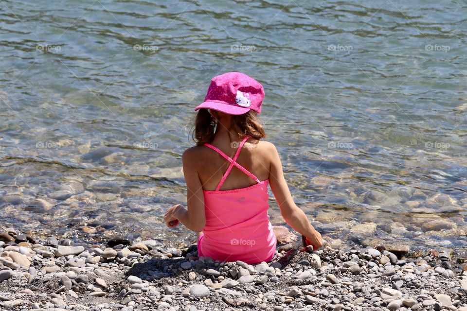 Little girl dressed in pink and pink baseball cap sitting beside the river 