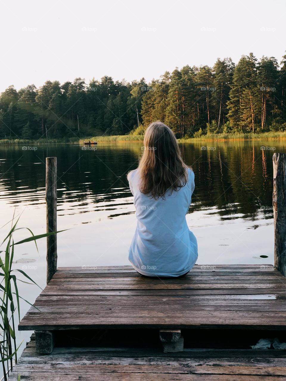 A young blonde girl sits on a wooden bridge by a lake on a sunny summer afternoon, back view 