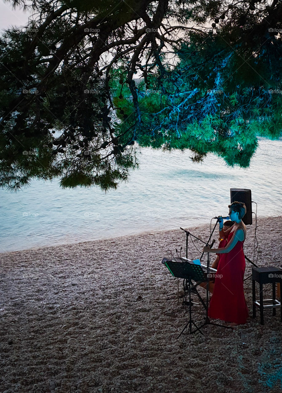 Summer evening on the warm south sea. A young girl in a red dress sings into a microphone on the beach on the shore of a calm sea under backlit pine trees