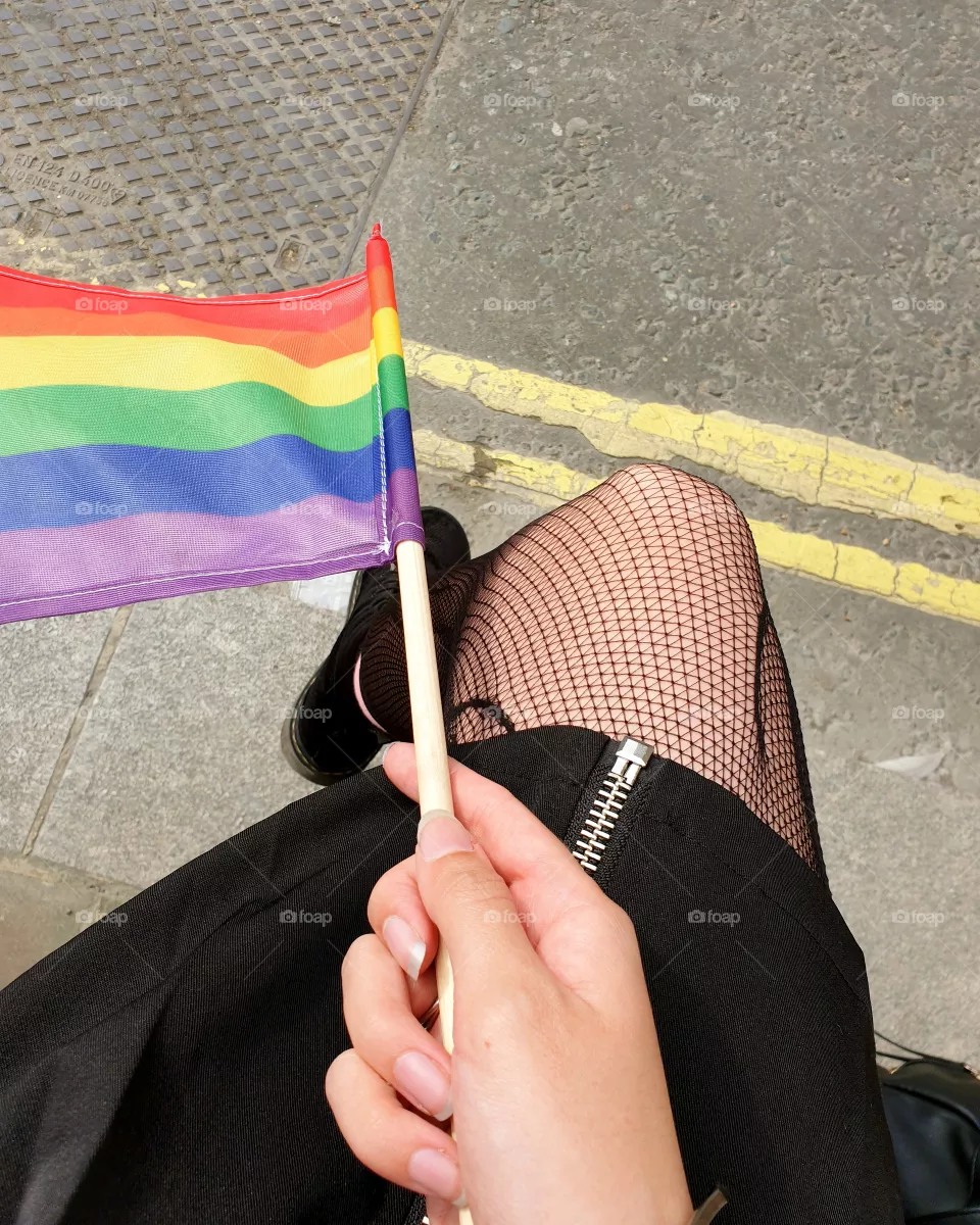 A hand holding a gay pride / rainbow flag on a street.