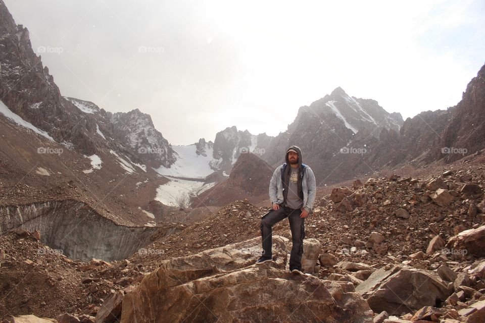 A man in a denim jacket with a hood is standing on a rock in the mountains on a glacier under falling snow