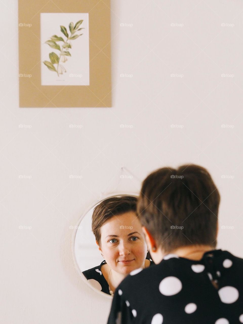 Portrait of a young beautiful woman with dark short hair looking into a round mirror in a bright room, portrait of woman