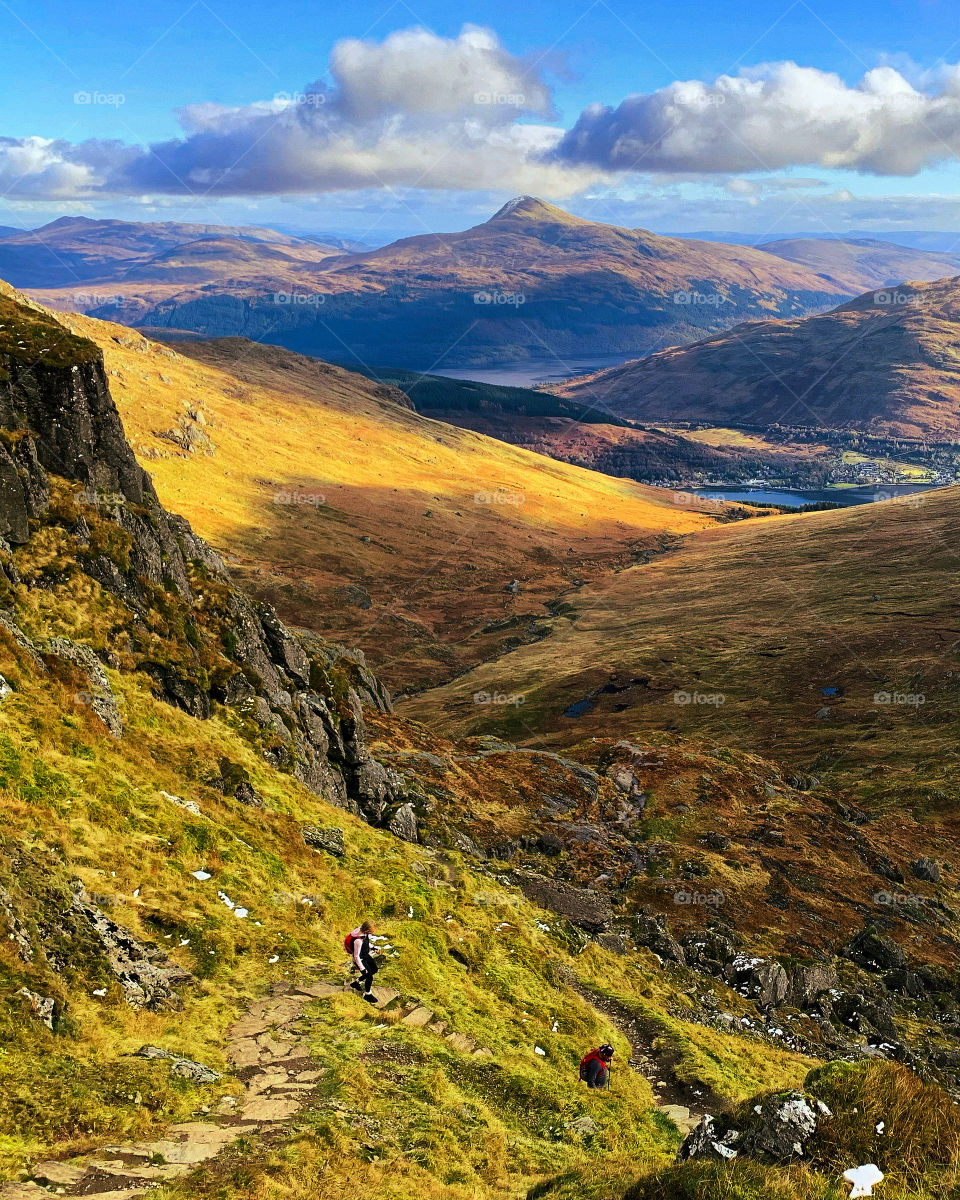 Sunny view of the beautiful landscape of the Scottish Highlands looking towards Ben Lomond. Hikers descending a trail on a popular mountain known as The Cobbler.
