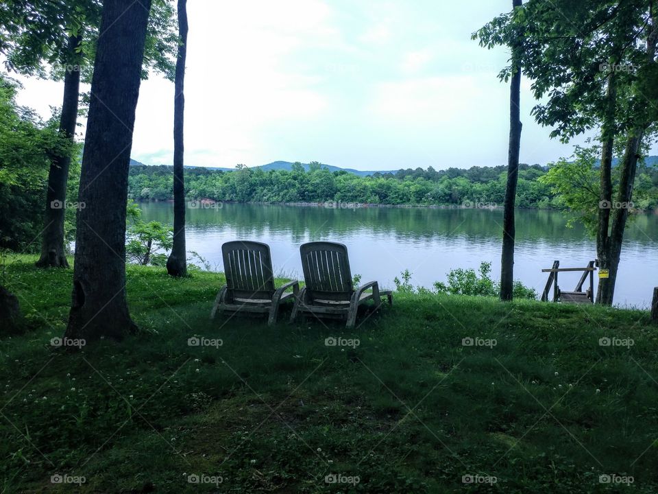 Two empty wooden lounge chairs facing a beautiful lake surrounded by greenery. Appears almost lonely or eerily but with a distinct sweetness and sense of natural beauty.