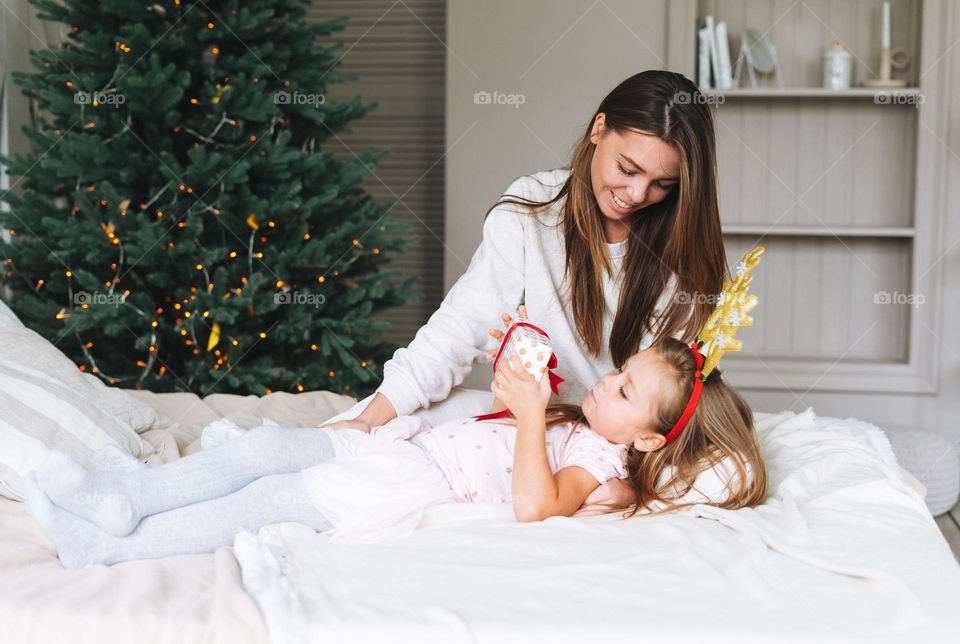 Cute little girl with mother on bed in room with Christmas tree. Portrait of child in the rim with deer horns in cottage house in christmas time, happy New Year. Happy family present christmas gift