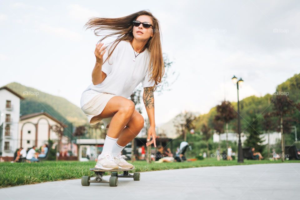 Slim young woman with long blonde hair in light sports clothes on longboard in outdoor skatepark at sunset time