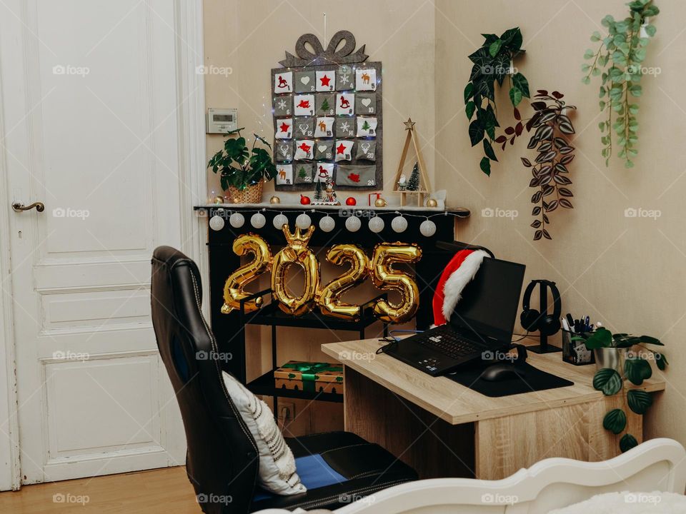 Beautiful desktop view with laptop, hanging flowers on the wall, wall-mounted Advent calendar and inflatable gold numbers 2025 on the black fireplace in the bedroom in the afternoon, close-up side view .