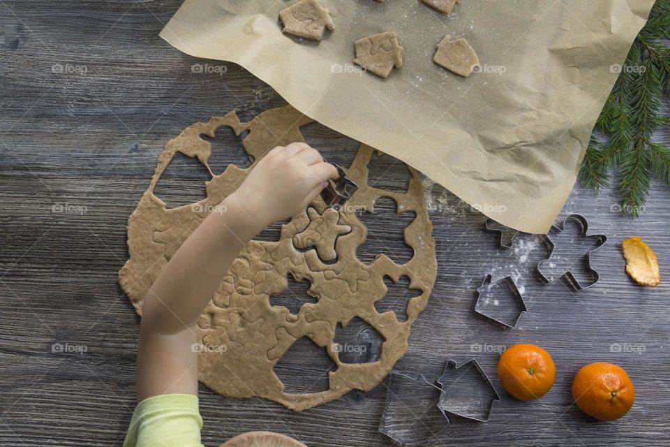 Little child helps cooking christmas ginger cookies on a wooden table with tangerines and green Christmas trees.