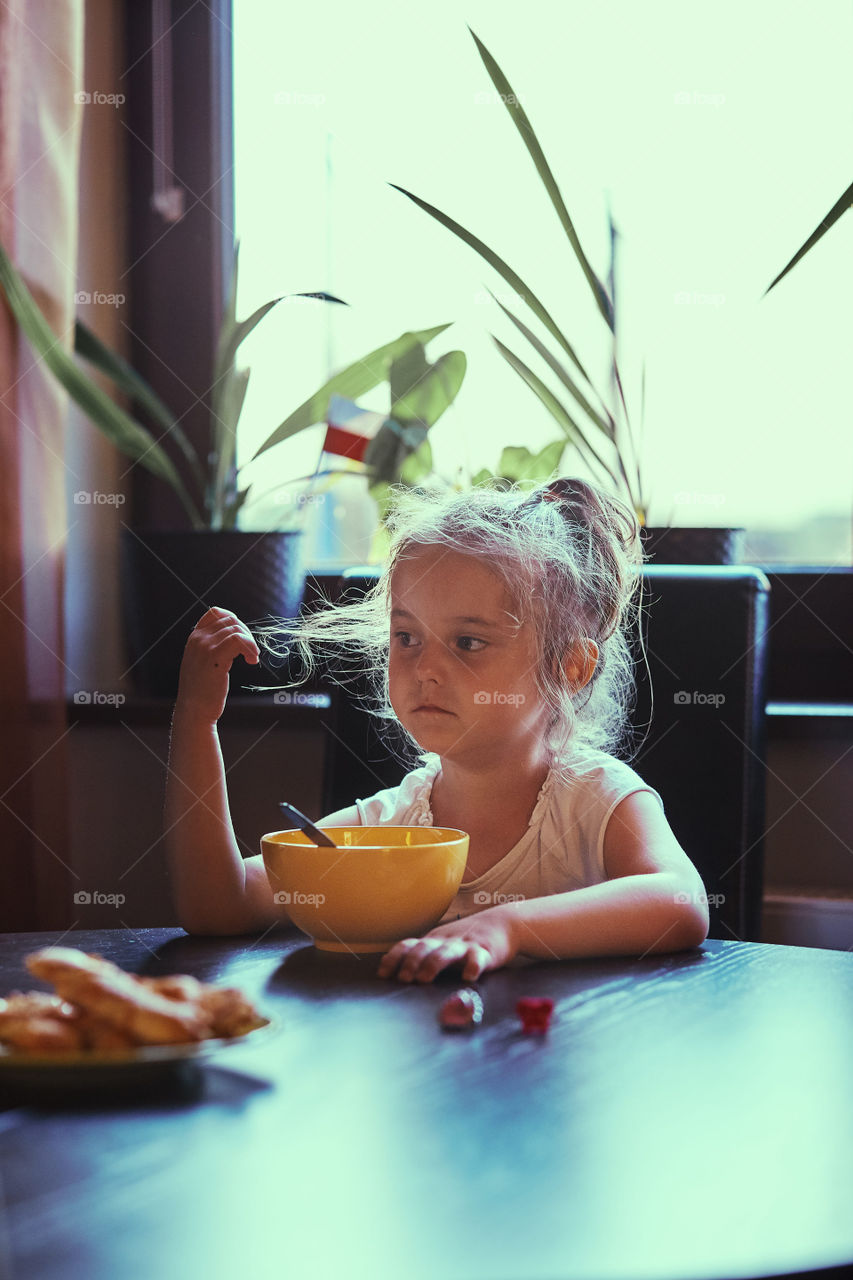 Little bored cute girl eating a breakfast, sitting by the table. Candid people, real moments, authentic situations