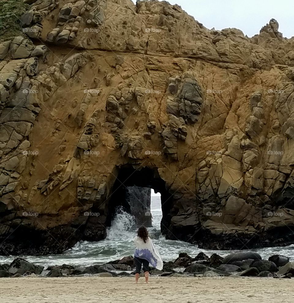 Women standing at an opening of a rock with ocean splashing through