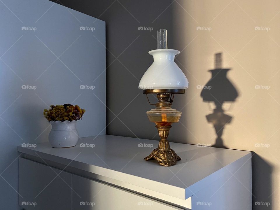 Beautiful vintage oil lamp with lamp shade made of white glass on a white cabinet. Shadow of the lamp on the wall. Modern interior design using an antique element.