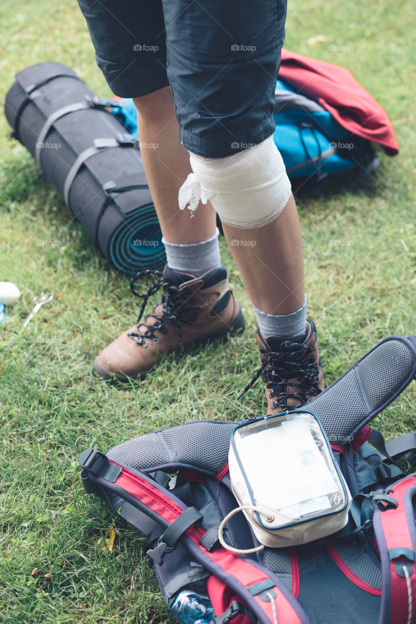 Woman injured in the accident on mountain hike. Dressing the wound on her knee with medicine in spray and gauze sitting on grass. Equipped with backpack wearing sportswear