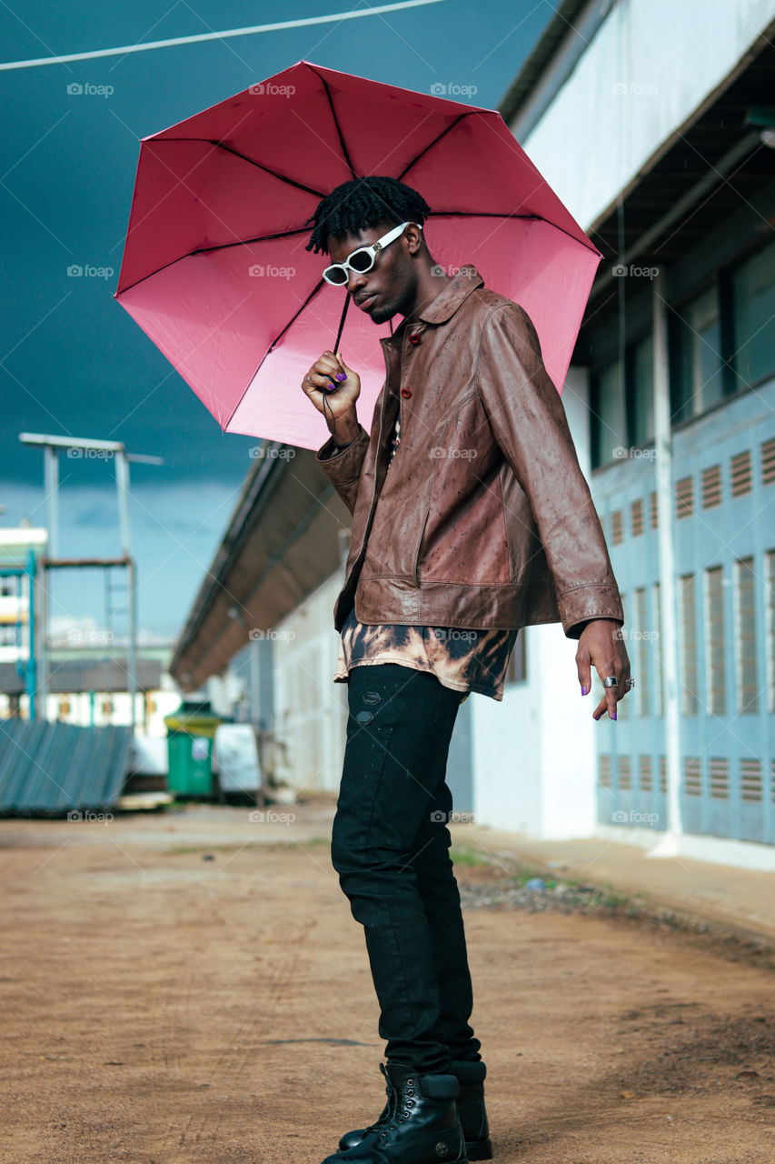 A boy holding a pink umbrella in the sun with dreadlocks hairstyle in brown jacket and a spectacle 