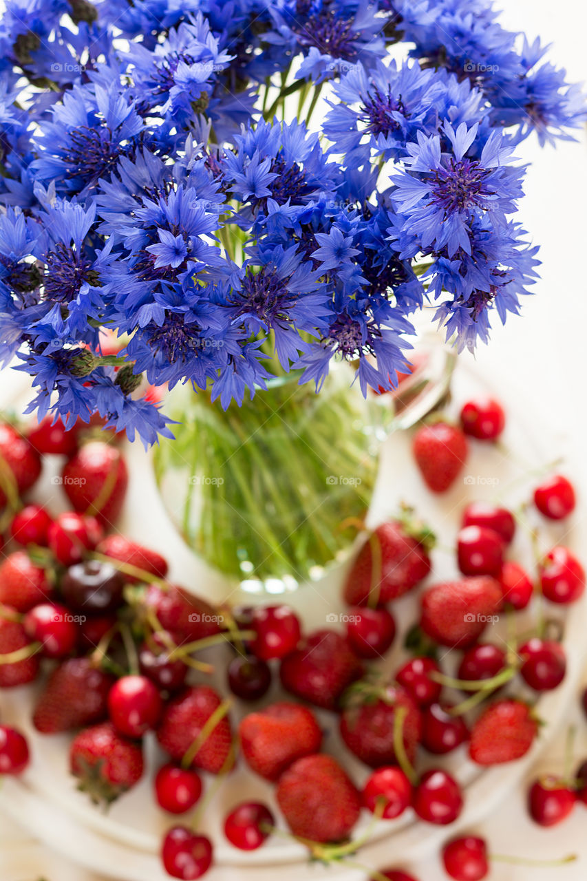 Cornflowers in vase, cherries and strawberries on white wooden background