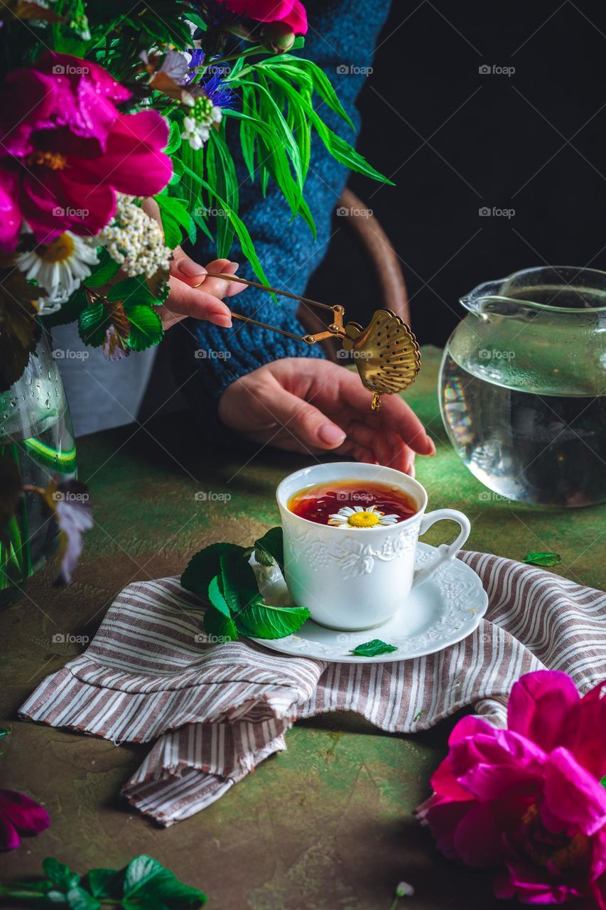 A girl in a blue sweater is preparing chamomile tea on the table, there is a bouquet of wildflowers nearby. Home sweet home. Herbal treatment and a natural, close to nature lifestyle, getting rid of stress. The joy of life