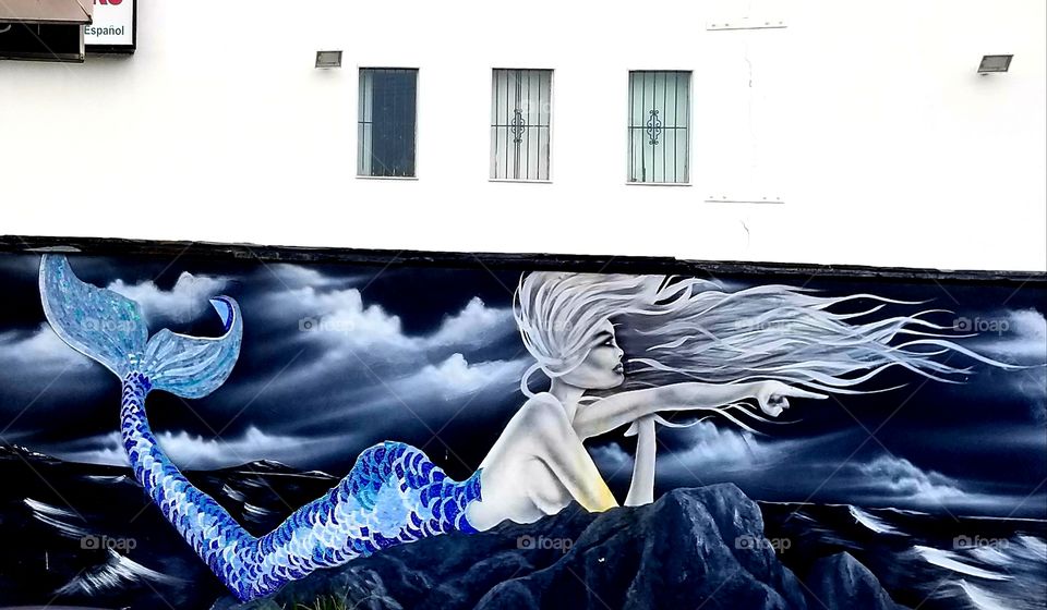 Mural of a mermaid/siren on the rocks. City wall. Blue painting.